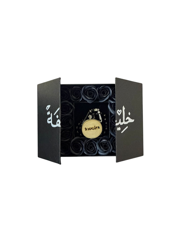 Black double-door gift box open, showing black preserved roses around a black masbaha, with the name engraved in white across the doors